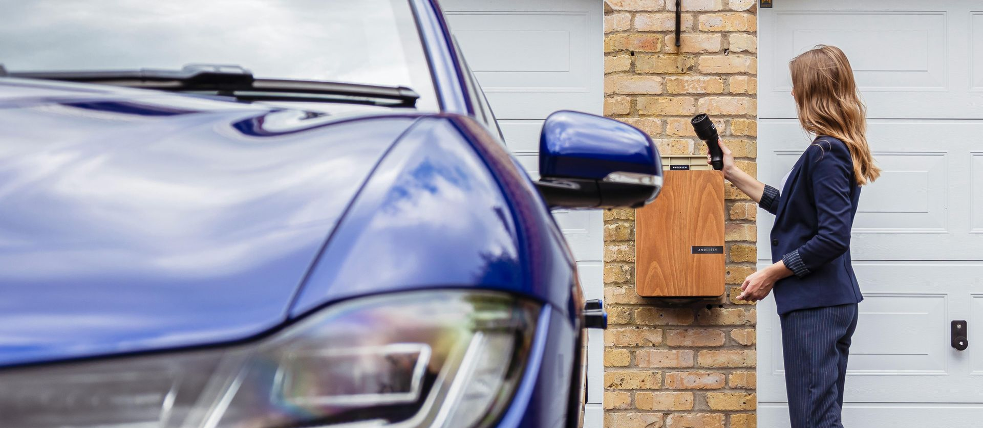 A woman in a blue suit checks a wooden mailbox near a blue car and white garage.