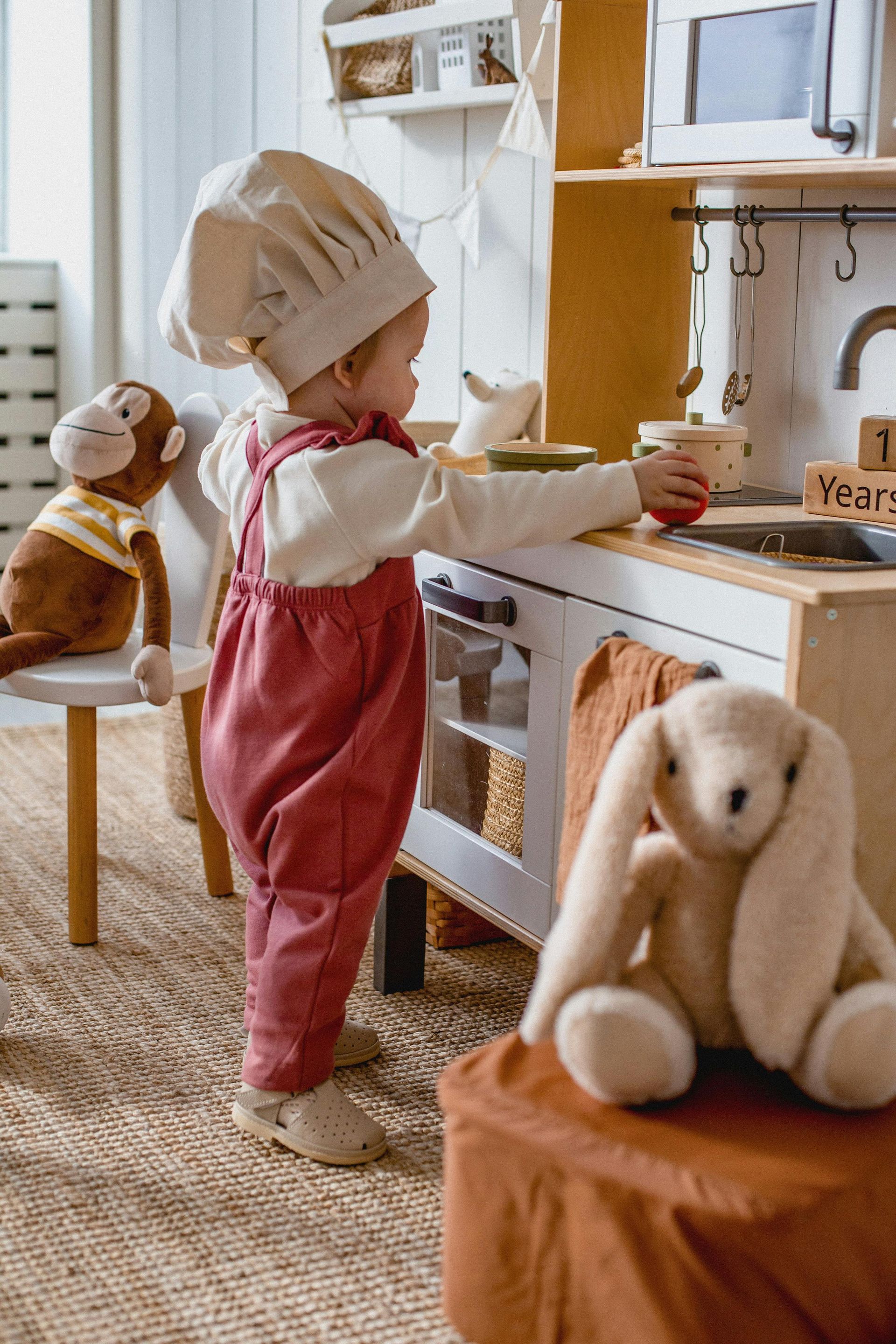 Child in chef hat and pink overalls plays at a toy kitchen, bunny and monkey plush toys nearby.