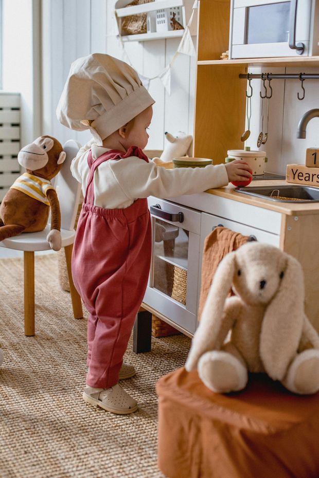 Child in chef hat and pink overalls plays at a toy kitchen, bunny and monkey plush toys nearby.