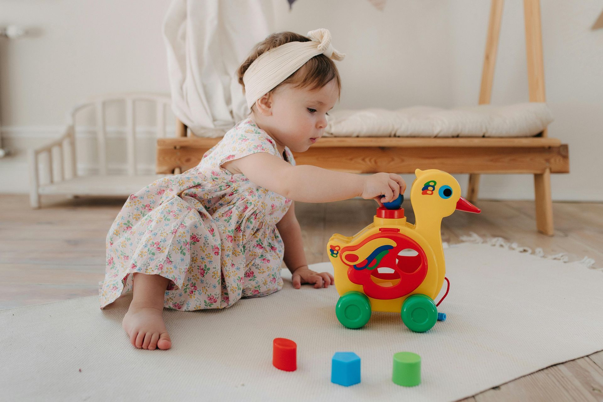A baby girl in a floral dress plays with a yellow duck toy and colorful blocks on a rug.