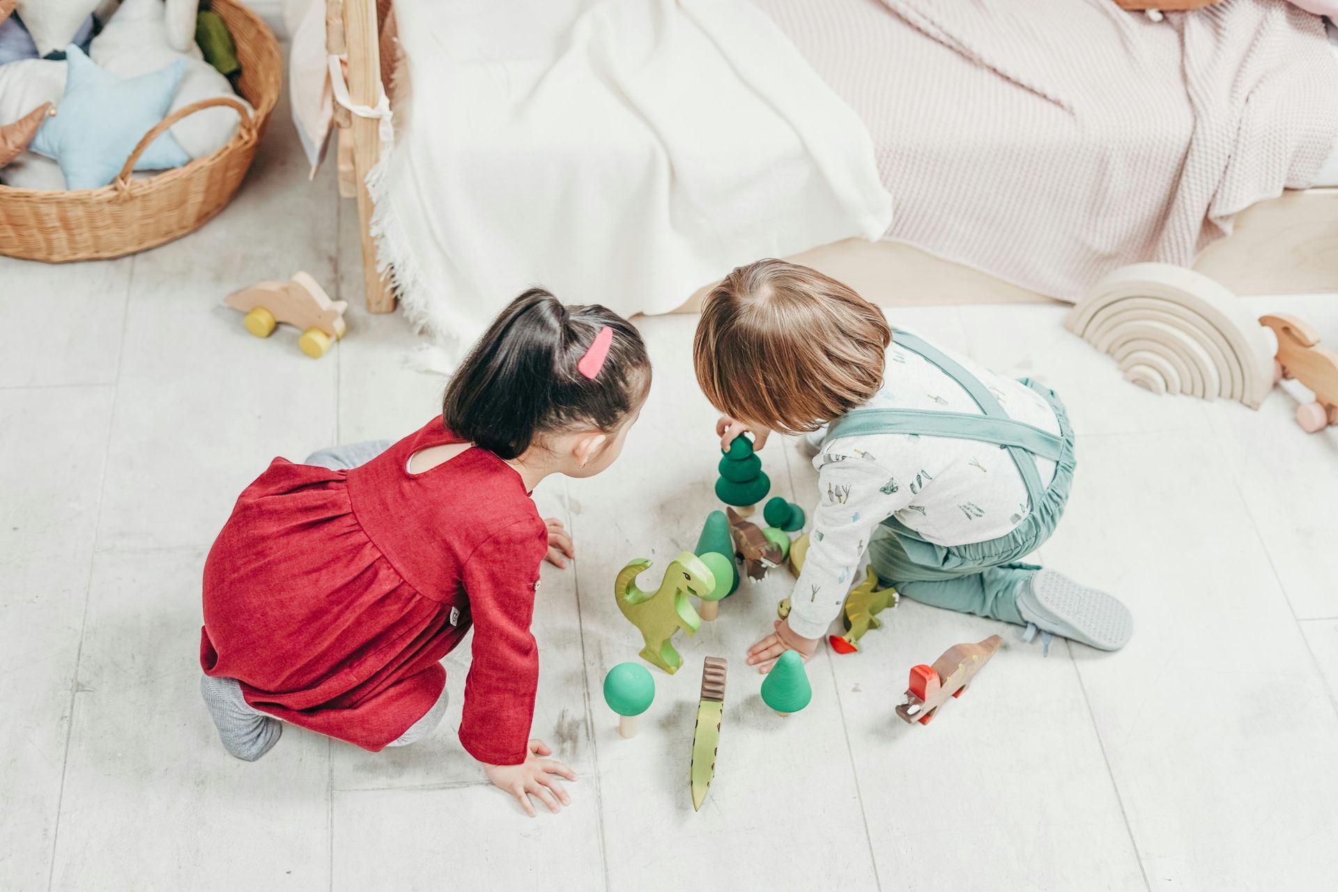 Two children play with wooden toys on a white floor. One wears red, the other blue overalls.