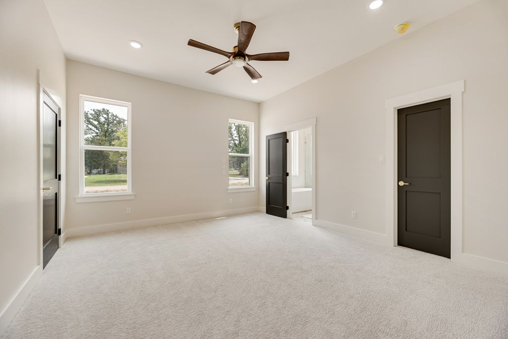 Spacious bedroom with neutral colors: white walls, gray carpet, brown ceiling fan, black doors, and windows.