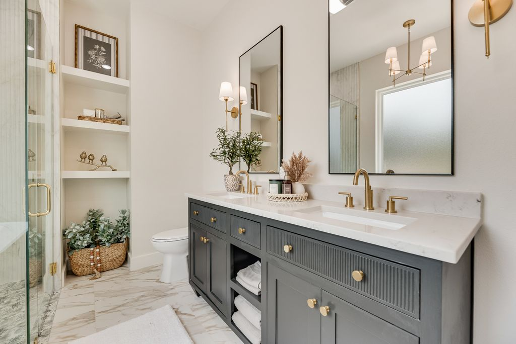 Bathroom with a dark gray vanity, gold fixtures, white countertops, and open shelving with decor.