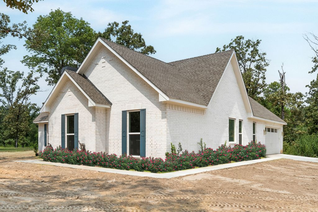 White brick house with blue shutters and brown roof; surrounded by bushes on a dirt lot.