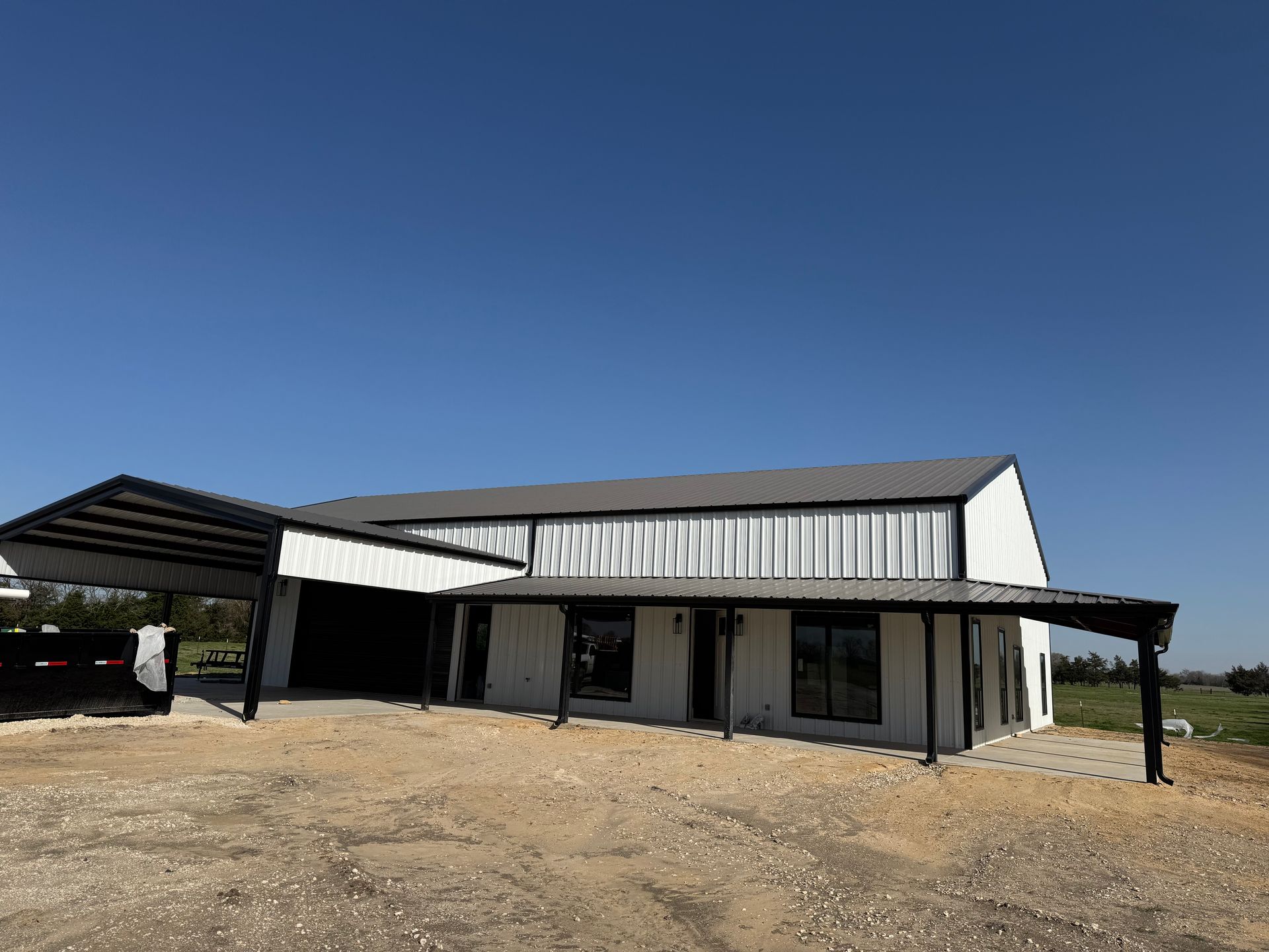 White barn with black accents and a carport under a blue sky.