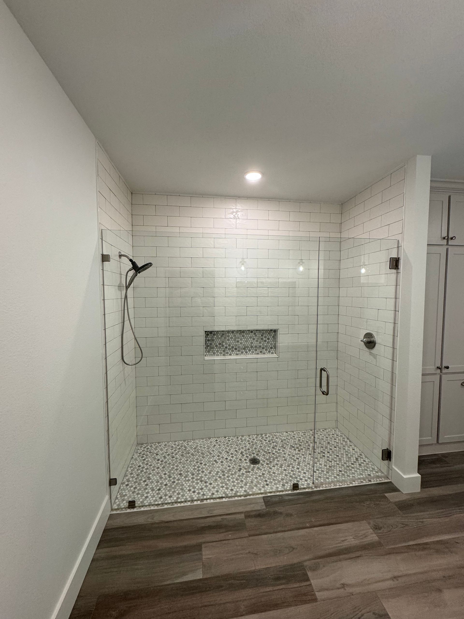 Modern walk-in shower with white subway tile walls, pebble floor, and glass doors. Dark fixtures and wood-look flooring.