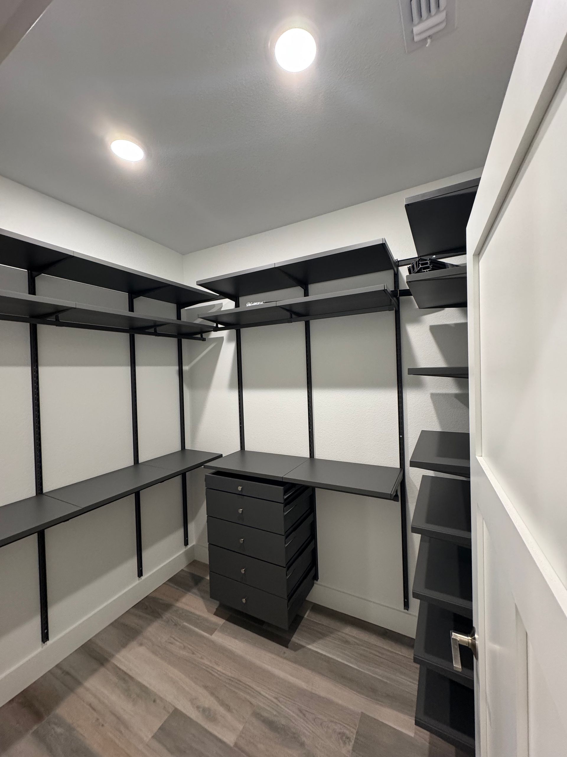 Empty walk-in closet with black shelving and drawers. White walls, wood-look floor, door on the right.