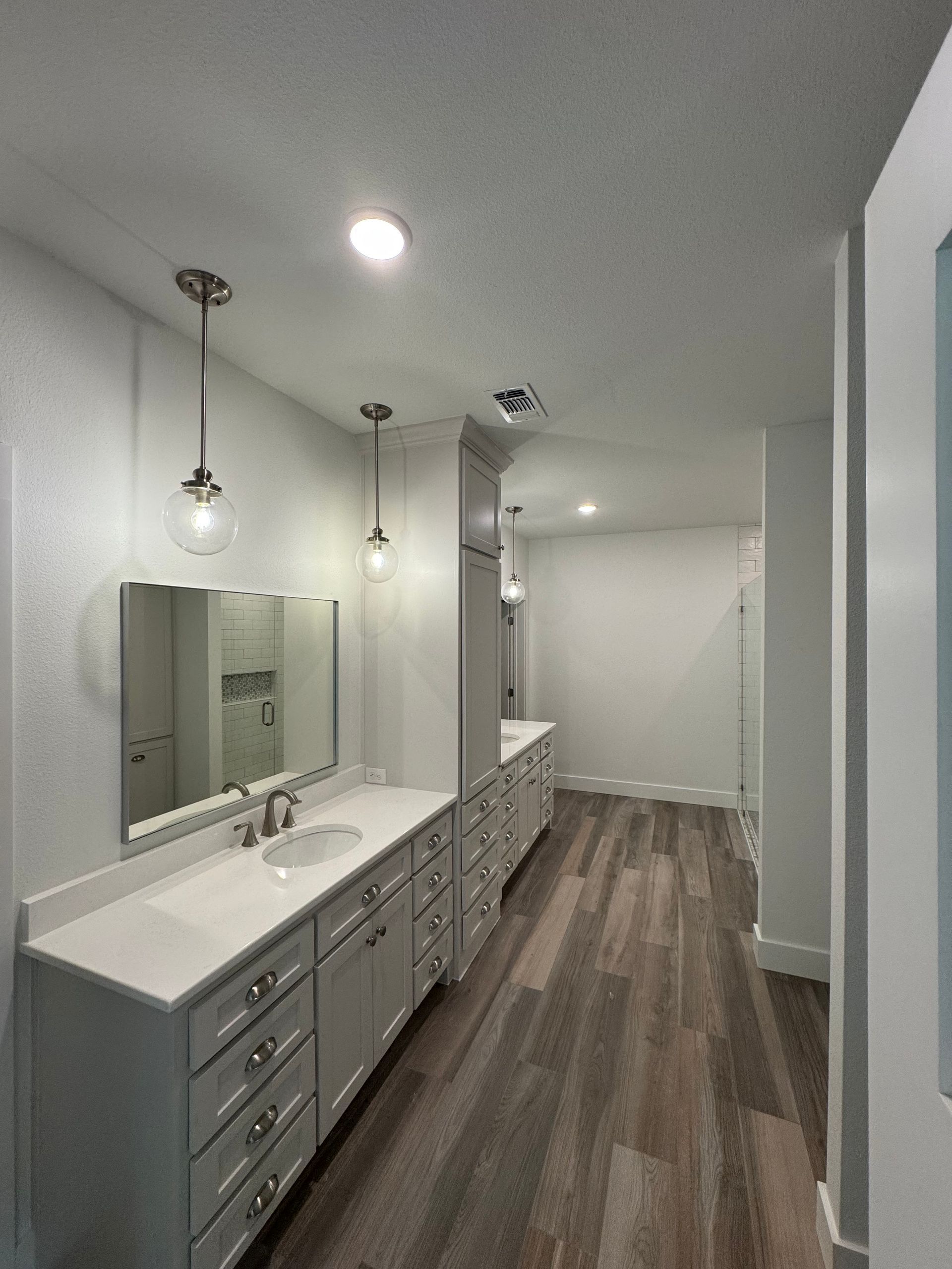 A long, white bathroom with gray cabinets, wood-look floor, and pendant lights.