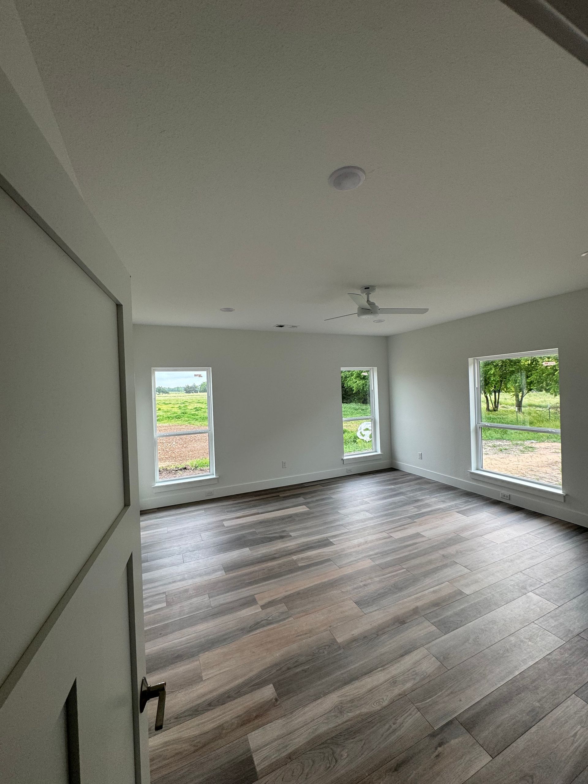 Empty room with wood-look floor, white walls, windows, and ceiling fan, overlooking a green field.