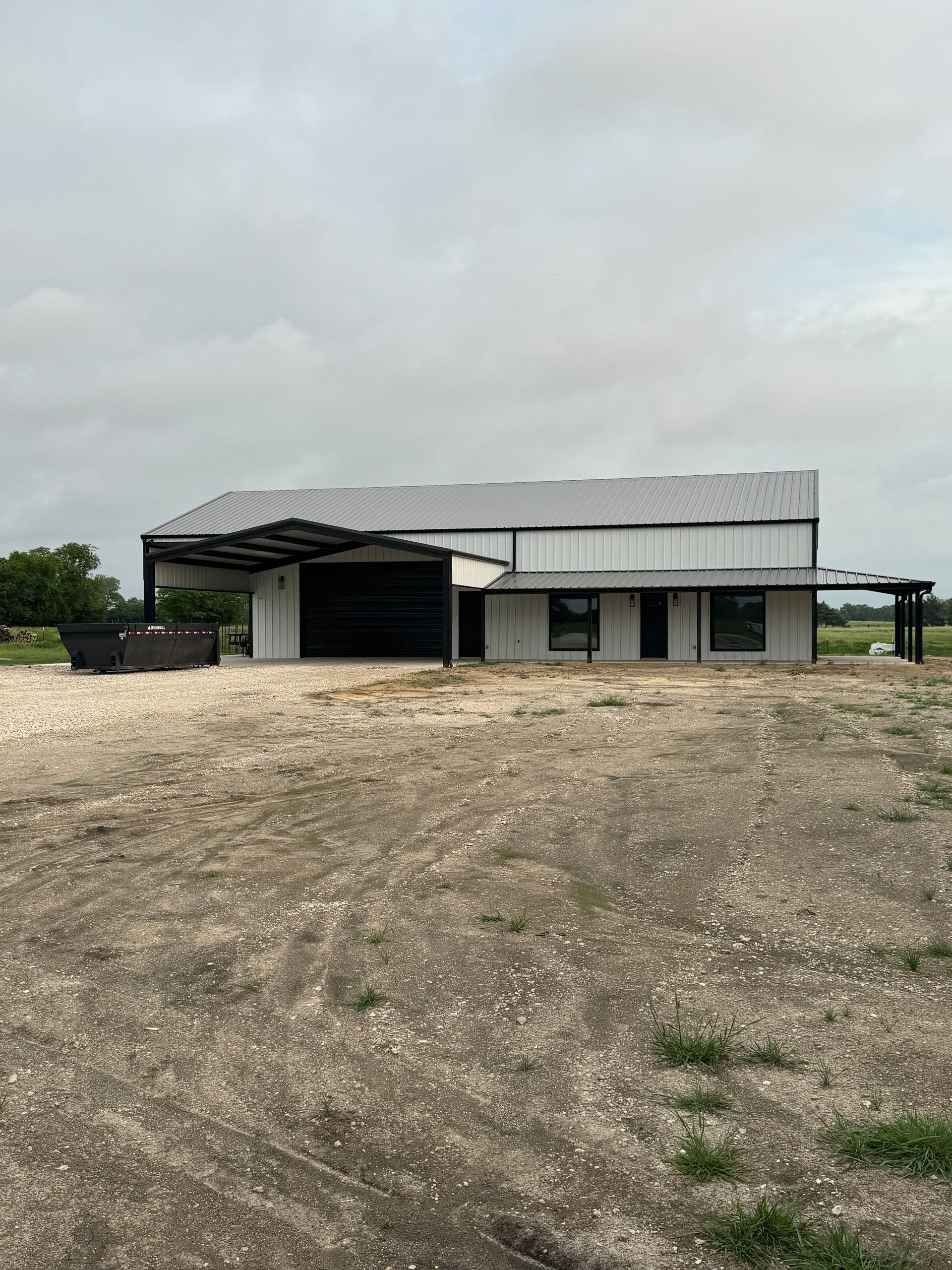 A large white barn-style building with a dark roof, set on a gravel and dirt lot under a cloudy sky.