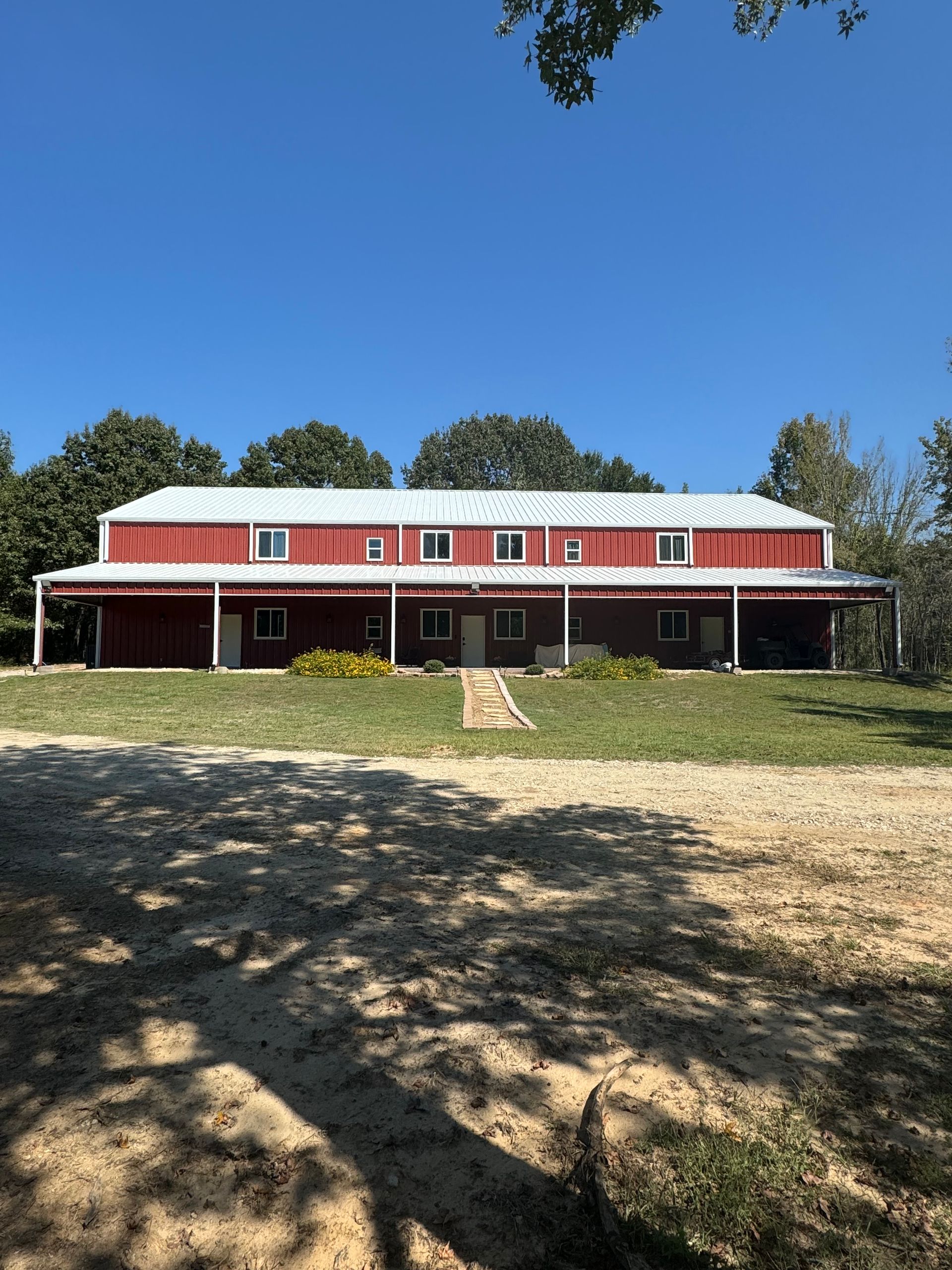 Red barn-like building with white trim, long porch, on a grassy area with a gravel driveway under a blue sky.
