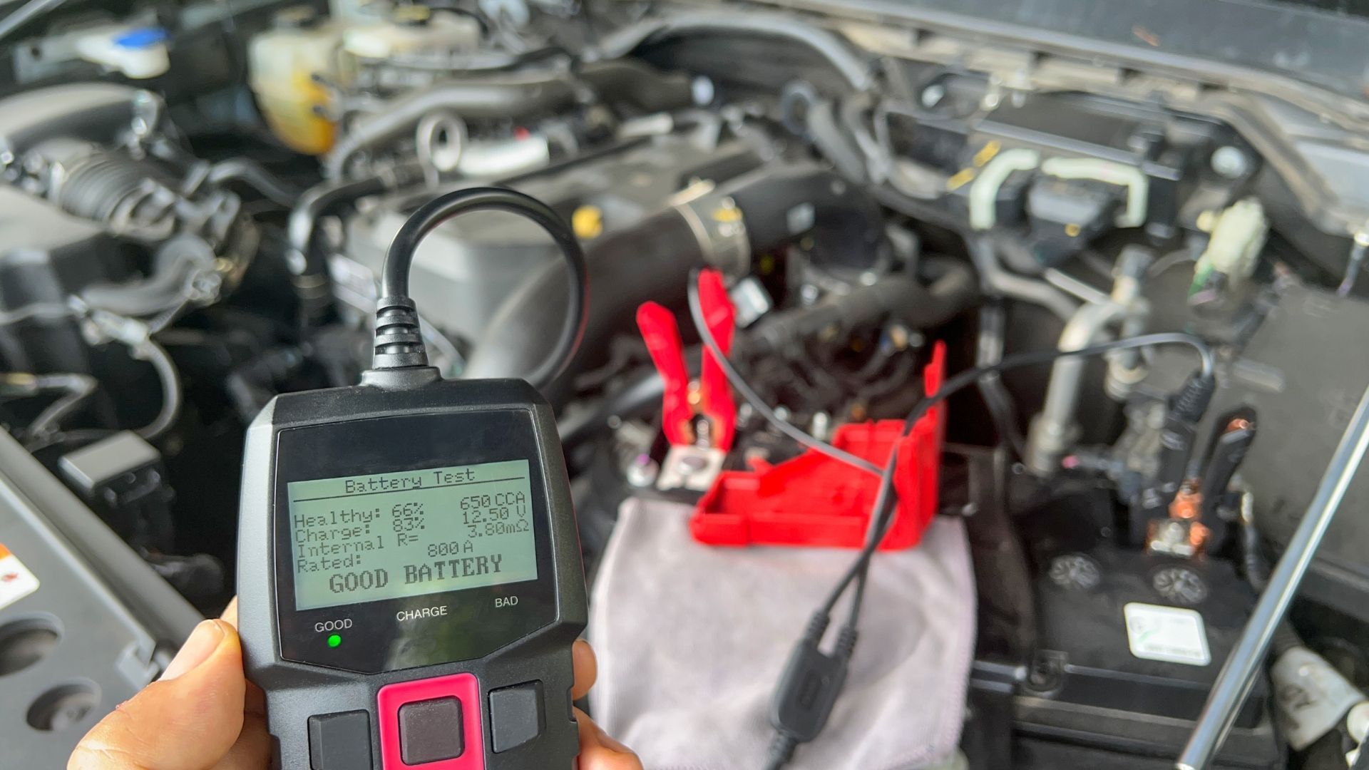 A car battery tester connected to a car battery in an engine bay. Screen shows test results, indicating 