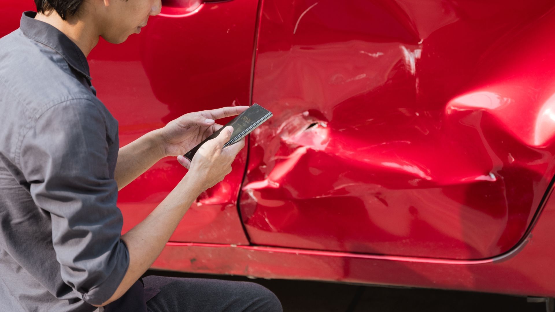 Person looking at car damage on a red car, using a phone.