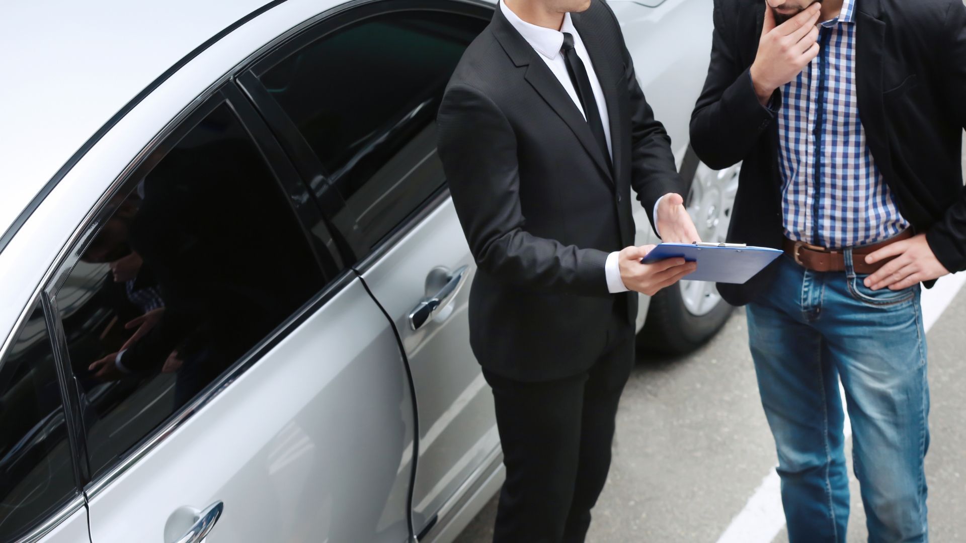 Man in suit shows paperwork to another man near a silver car.