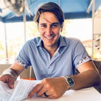 Man smiling, holding a paper, wearing a blue shirt and a watch, sitting outdoors.