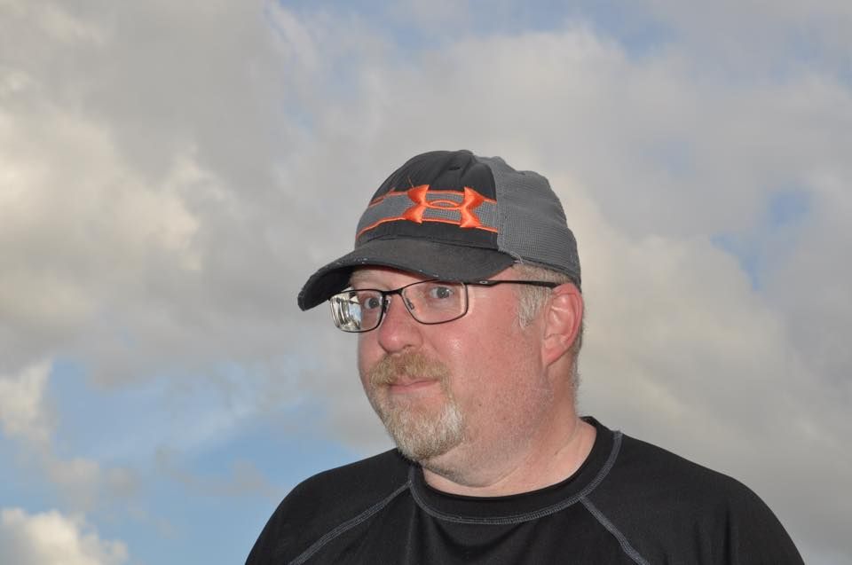 Man wearing glasses and cap, looking to the side against a cloudy sky.
