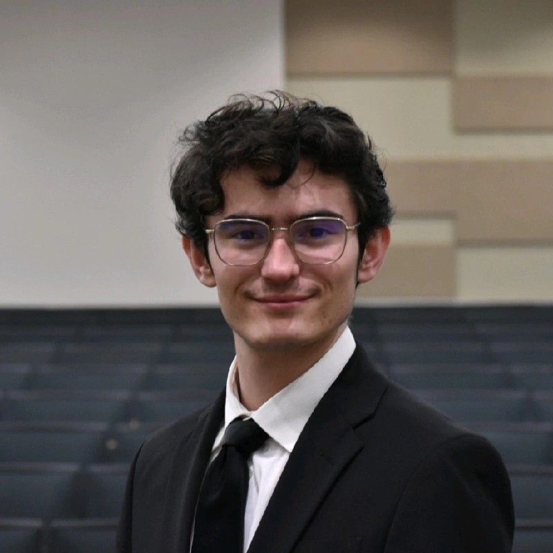 A young man with glasses, wearing a black suit and tie, smiles at the camera in a room with a stage.