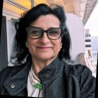 Woman with black hair and glasses smiles outdoors. She wears a black jacket and pearl necklace.