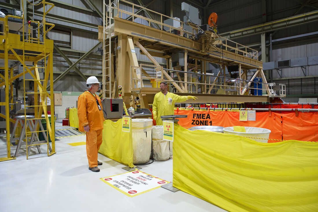 Two workers in protective suits stand near a machine inside a large industrial building. The setting suggests a nuclear facility.