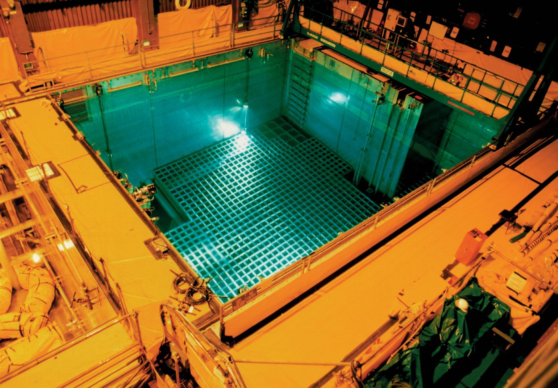 A deep blue nuclear reactor cooling pool glowing underwater. Tall rectangular racks hold spent nuclear fuel rods.