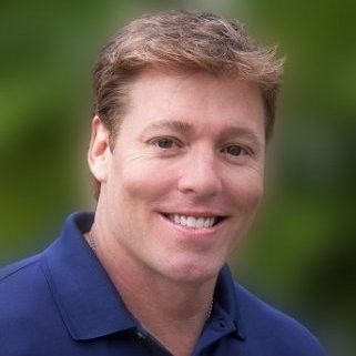 Smiling man with short, light brown hair wearing a blue polo shirt, against a blurred green background.