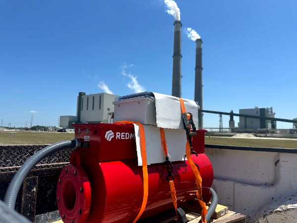 A red industrial machine is strapped down outdoors with power plant smokestacks in the background.