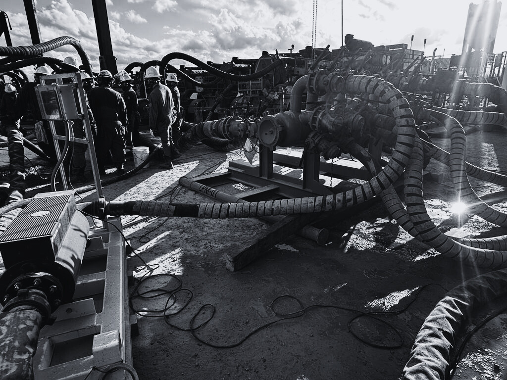 Workers near industrial equipment, potentially oil or gas infrastructure, with many hoses and pipes on a construction site.