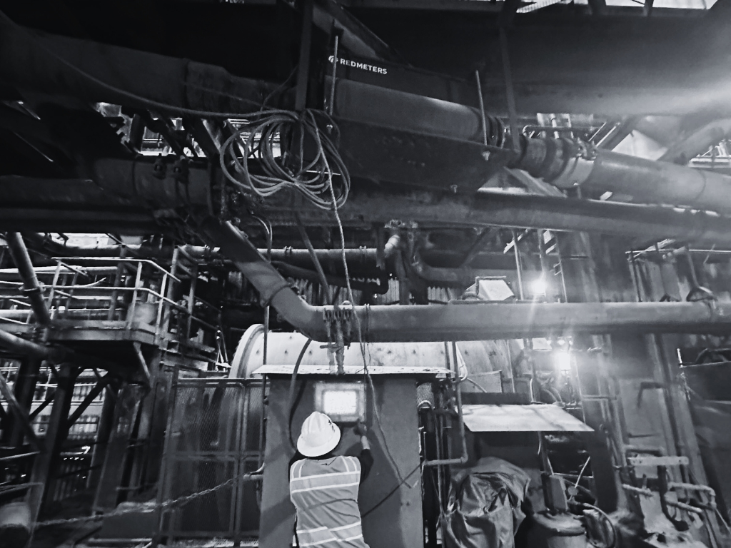 A worker in a hard hat and safety vest inspects machinery in an industrial setting, surrounded by pipes and wires. Black and white.
