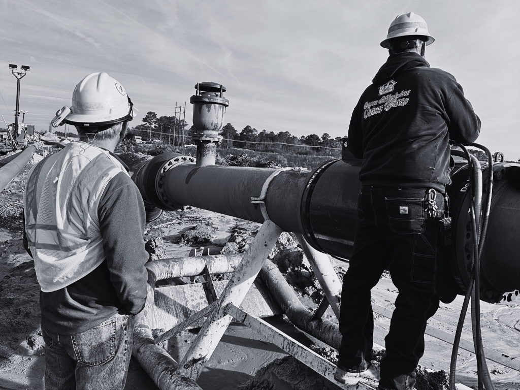 Two construction workers in hard hats examine a large pipe outdoors. One worker stands near the pipe, while the other works on it from a small platform.