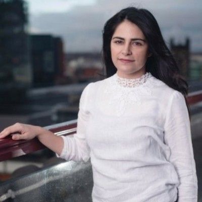 Woman with dark hair in a white blouse, smiling slightly, leaning on a railing overlooking a city.