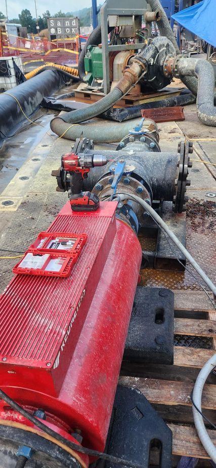 A red meter industrial pump connected to pipes on a wooden platform. There are tools and other equipment nearby, possibly a construction site.