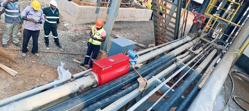 Workers in hard hats and safety vests examine a red electrical box on a series of pipes. Industrial setting with metal structures and ground visible.