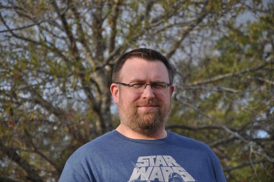 Man with glasses and beard wearing a Star Wars t-shirt, smiling in front of a tree.