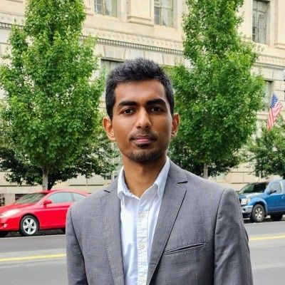 A man in a grey suit and white shirt stands in front of a building. He smiles slightly, with a red car and blue SUV in the background.
