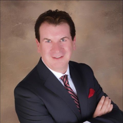 Man in a suit smiling, arms crossed, red pocket square, and a striped tie. Background is a neutral brown.