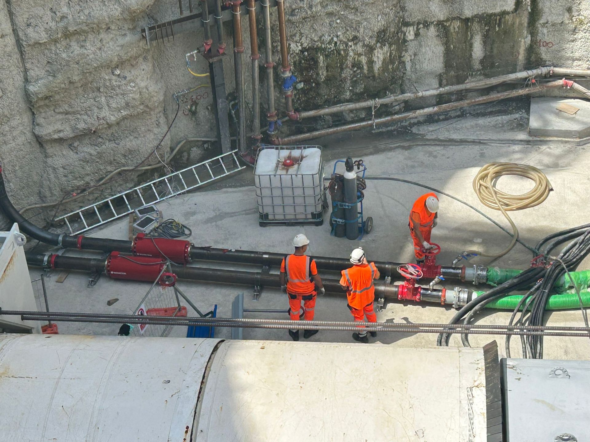 Construction workers in orange vests and white helmets are working on pipes in an outdoor concrete setting.