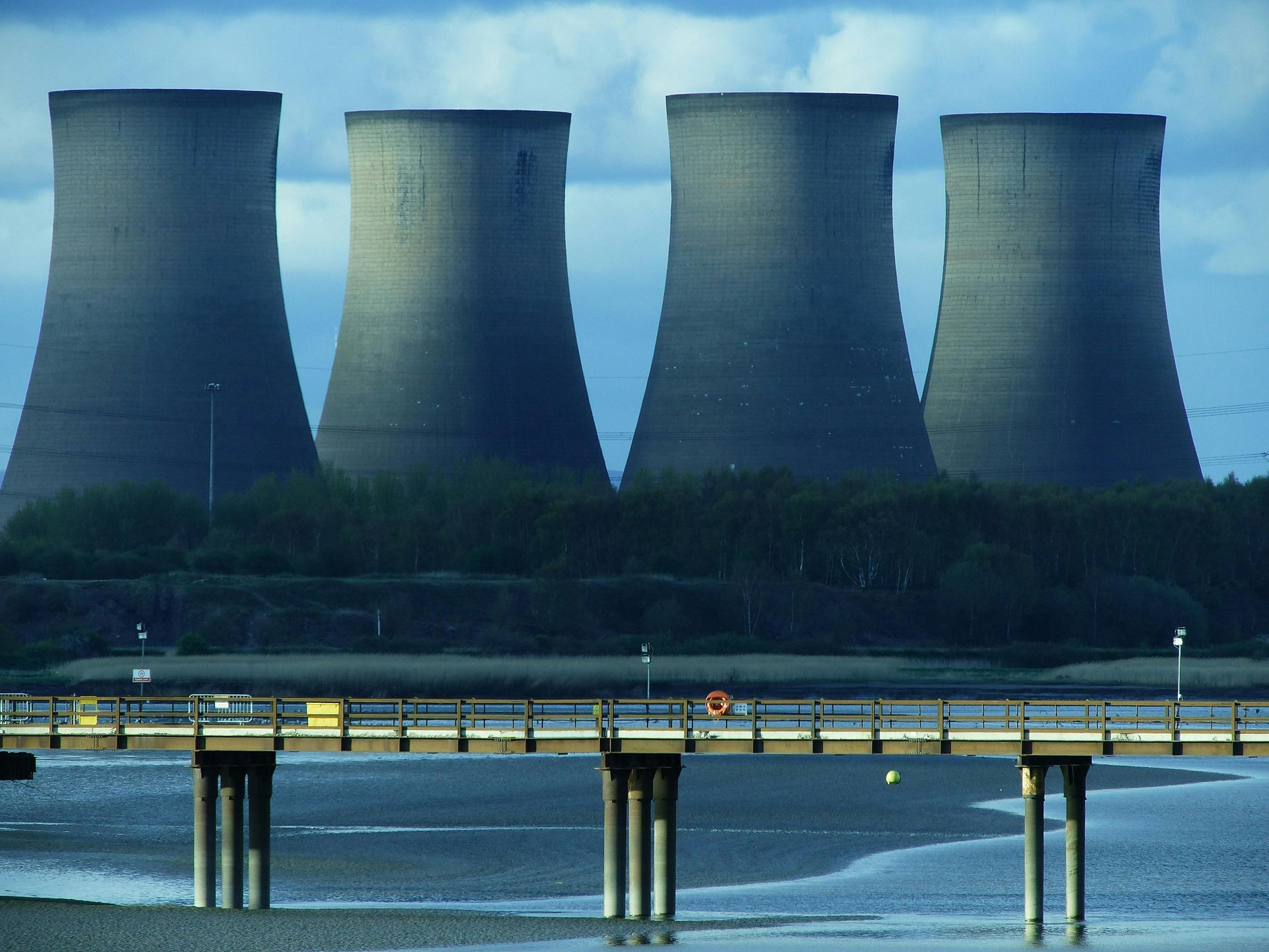 Four large cooling towers stand against a blue sky, with a pier in the foreground.