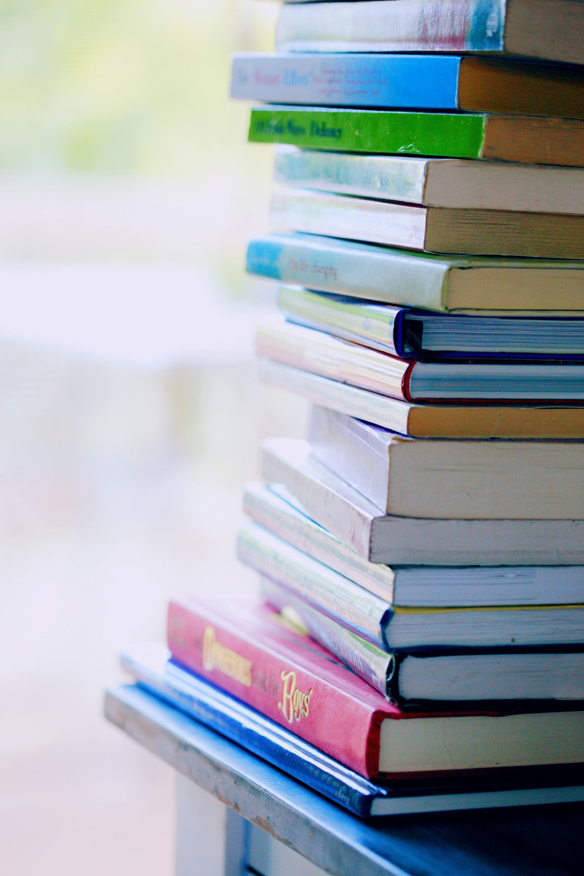 Stack of books on a table, varying colors and sizes. Soft sunlight in the background.