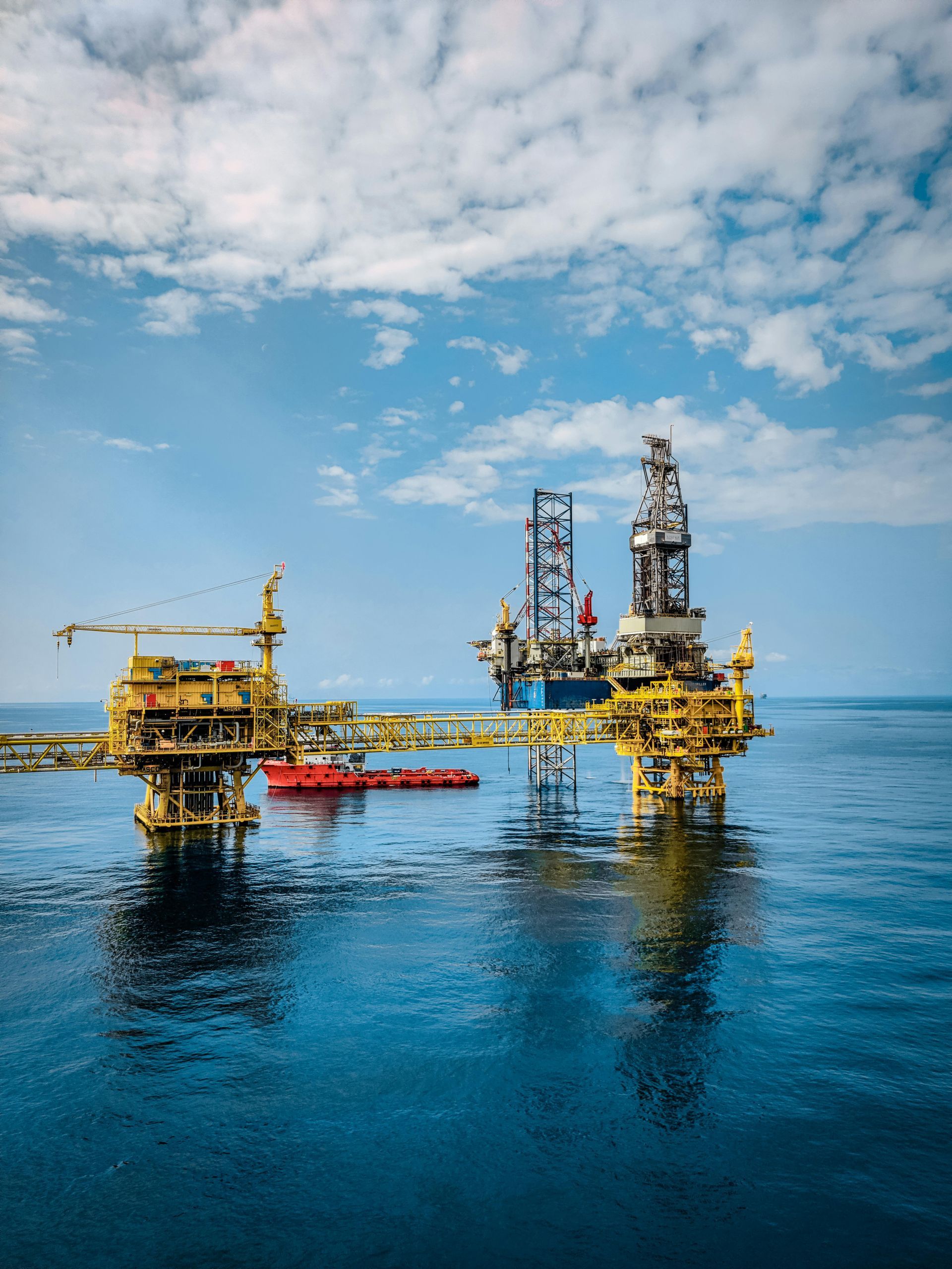 Offshore oil platforms in blue water under a partly cloudy sky. Yellow and red structures with a blue drilling rig in the background.