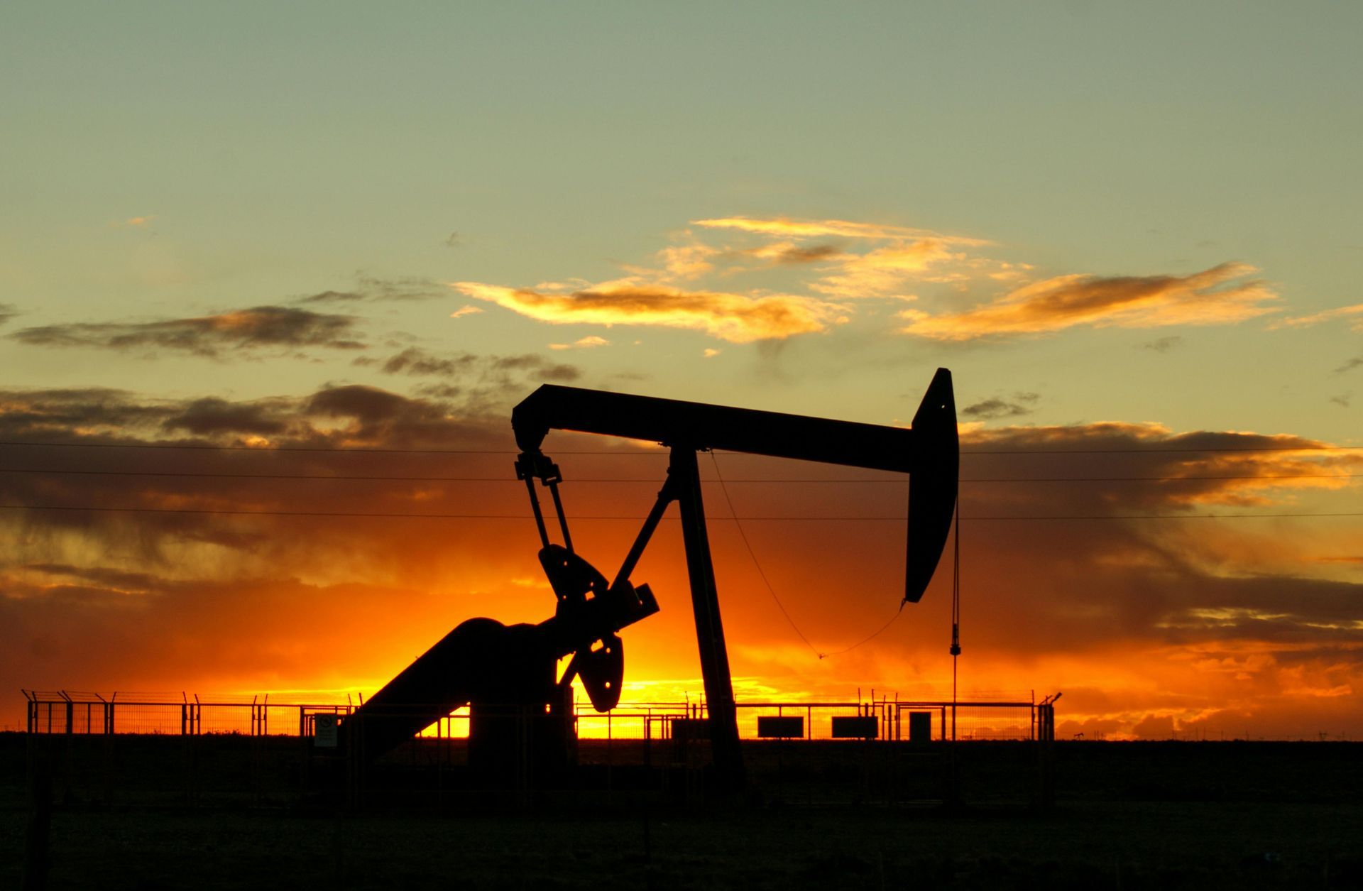 Silhouette of an oil pump jack against a sunset sky with orange and yellow hues.