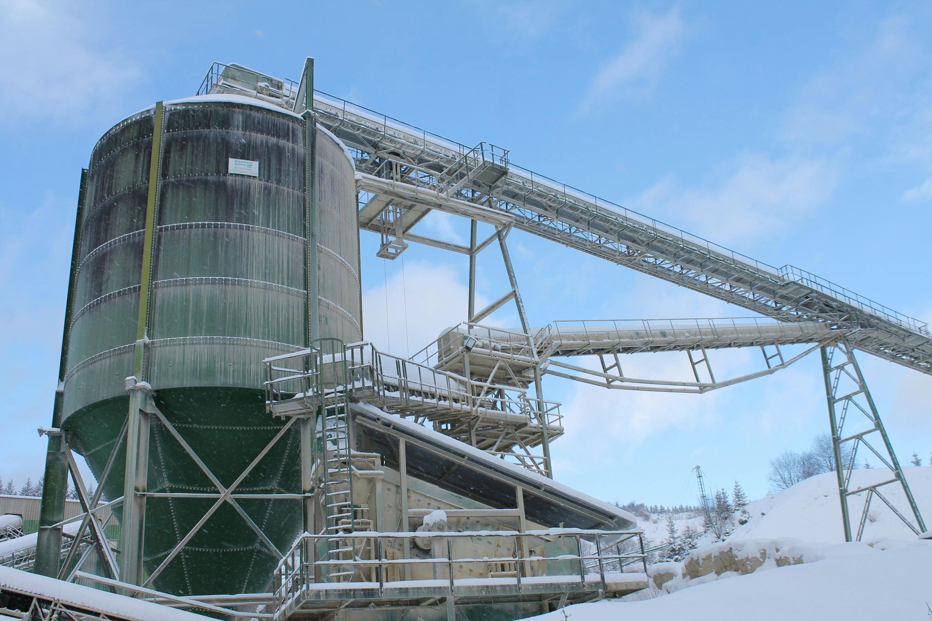 Snow-covered industrial plant with a large silo and conveyor belts against a blue sky.
