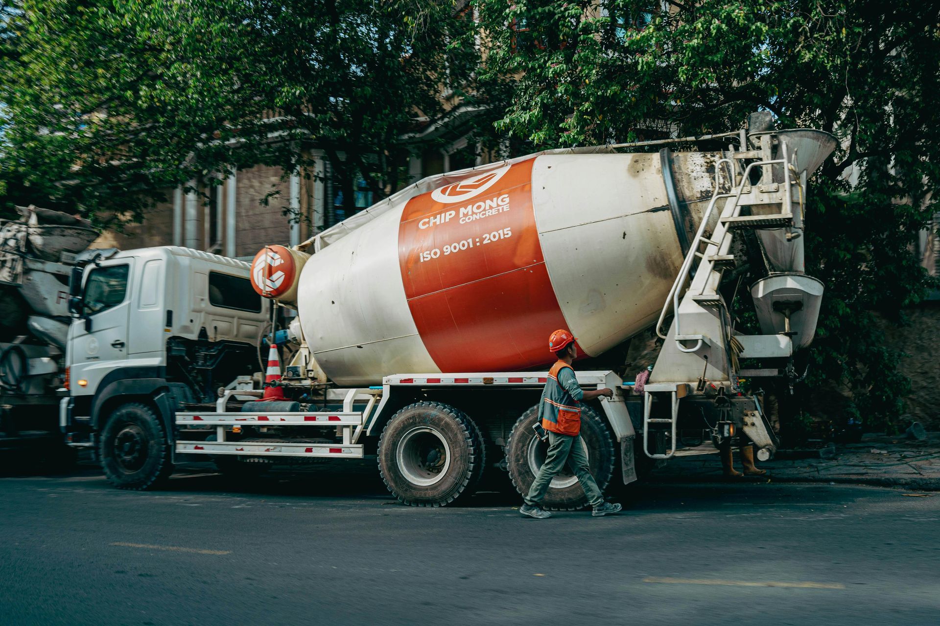 Cement truck on a city street. A worker in a hard hat stands near the truck's side, with buildings and trees in the background.