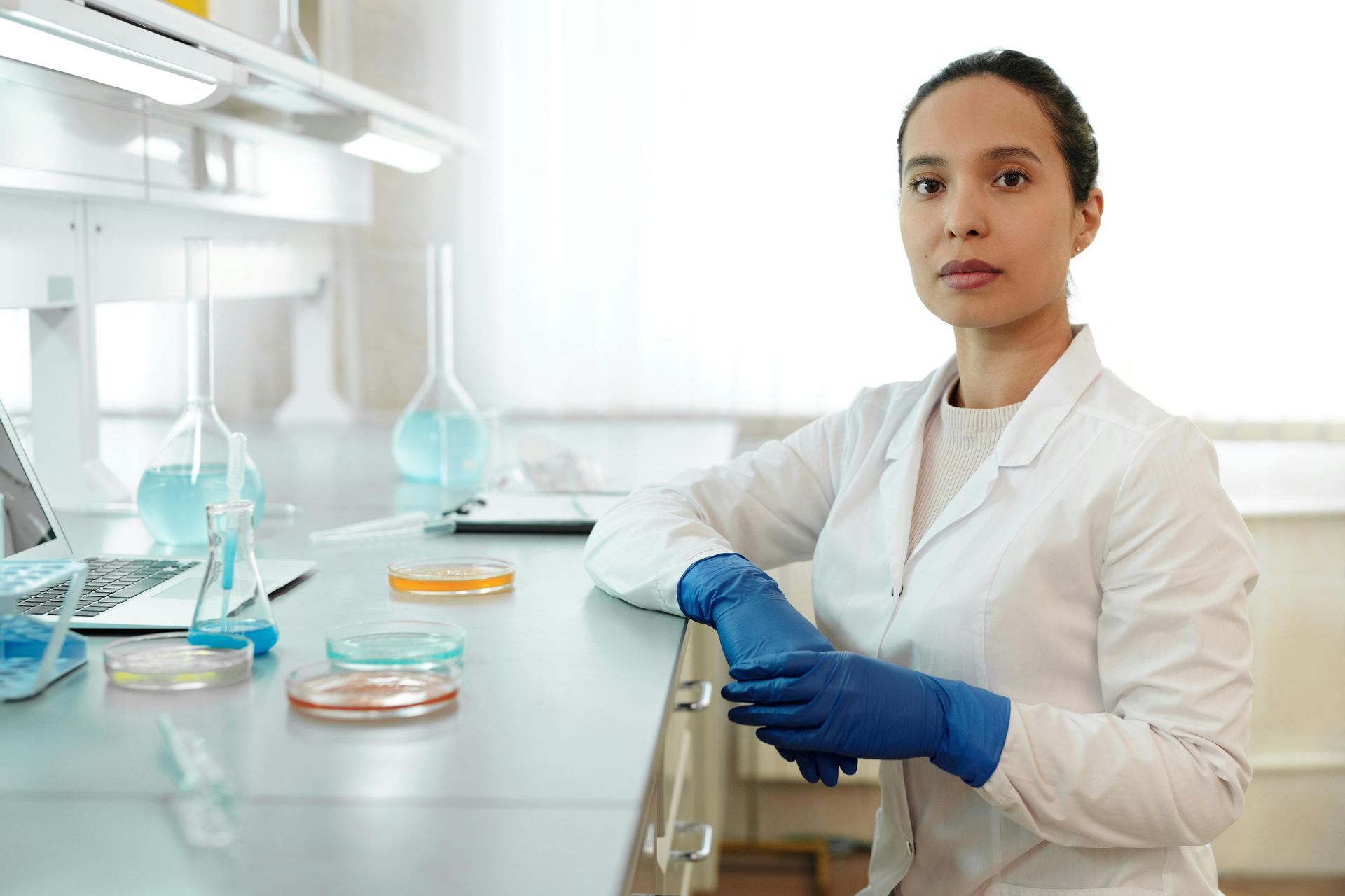 A woman in a lab coat and gloves in a lab, gazing at the viewer. Petri dishes and lab equipment are on the counter.