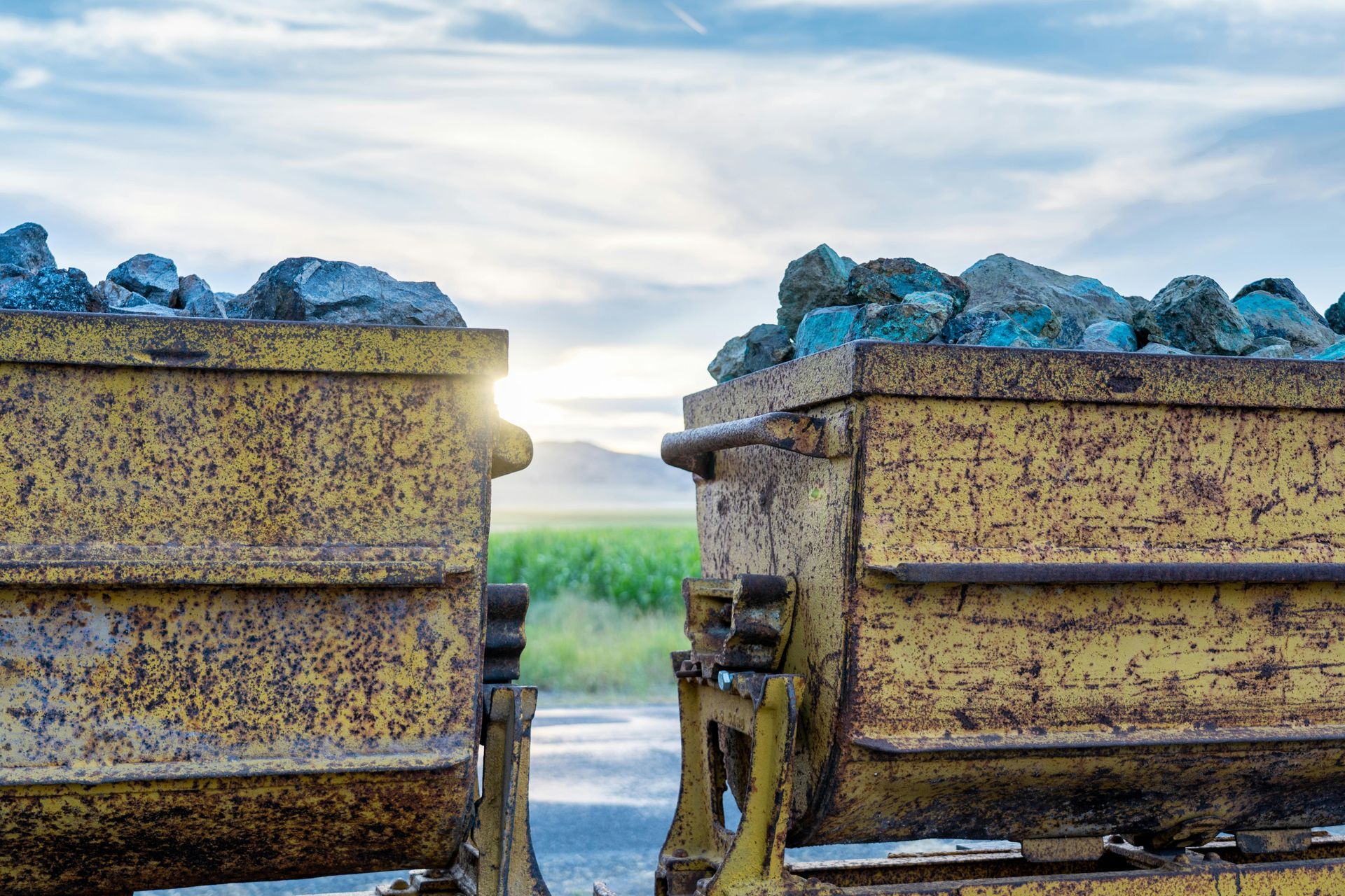 Two rusty yellow mining carts filled with rocks, set against a green field and a sky with the sun peeking through.