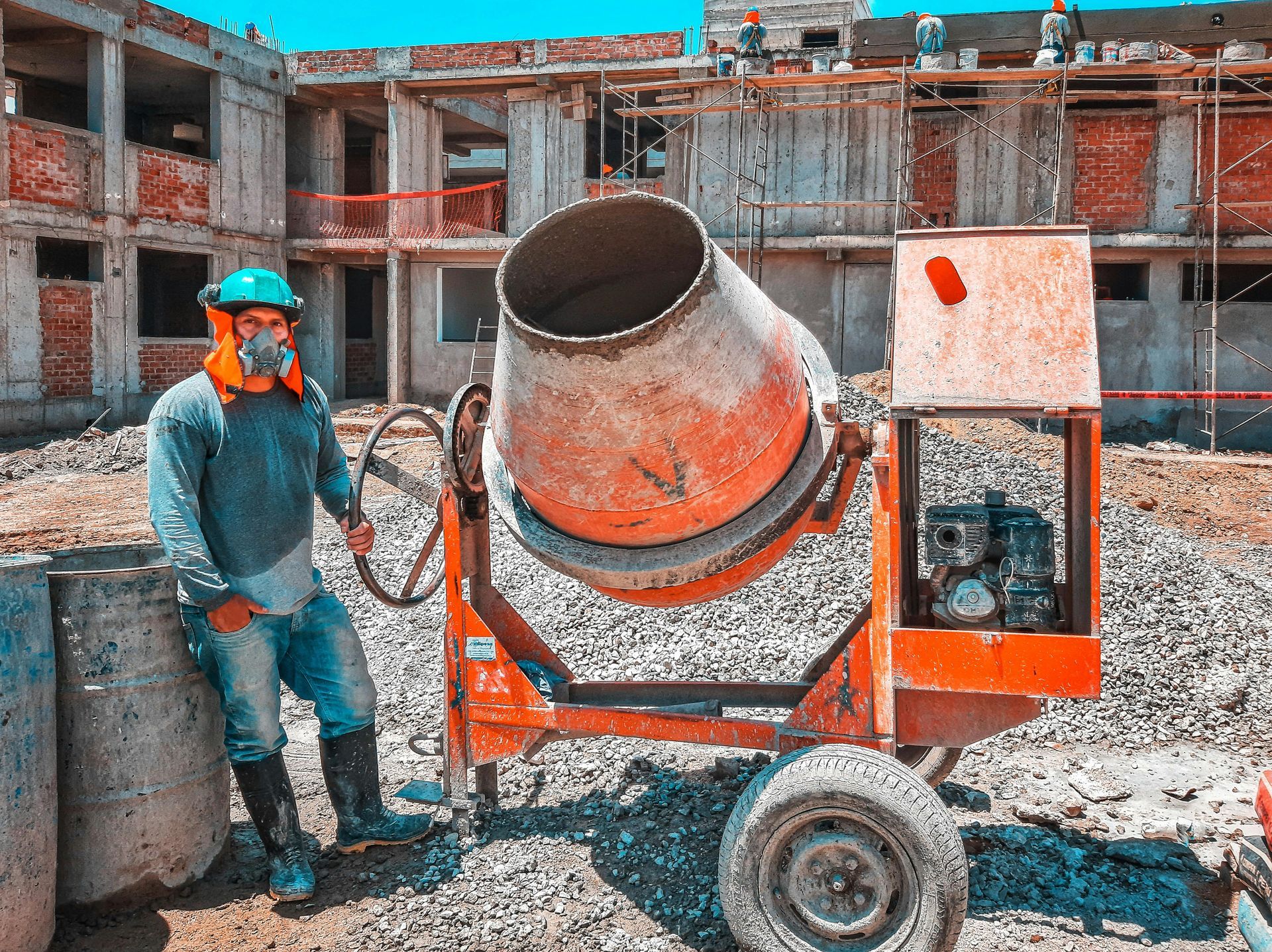 Construction worker beside an orange cement mixer at a building site. Worker wears protective gear, leaning against a concrete cylinder.