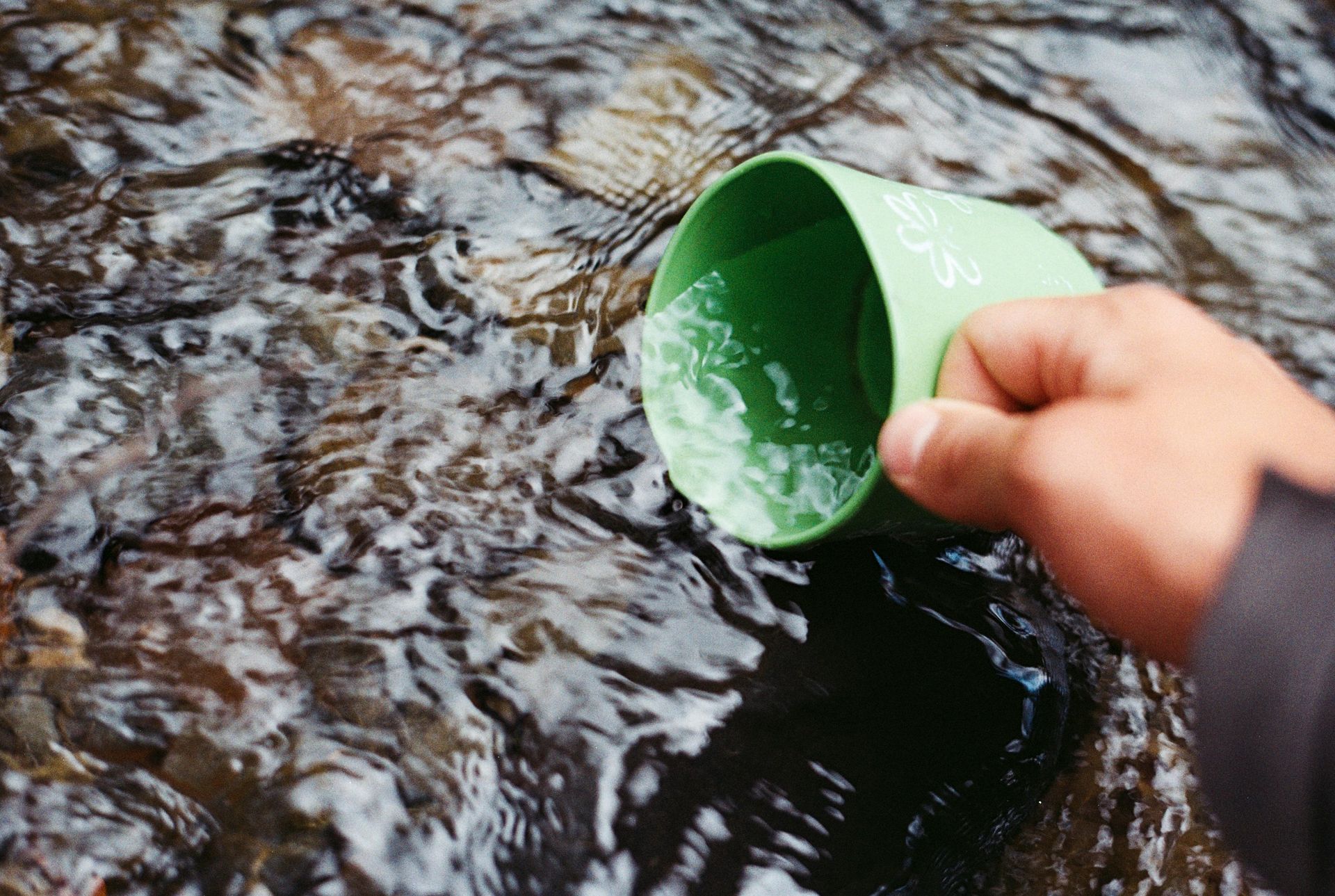 Person's hand scoops water with a green cup from a flowing stream.