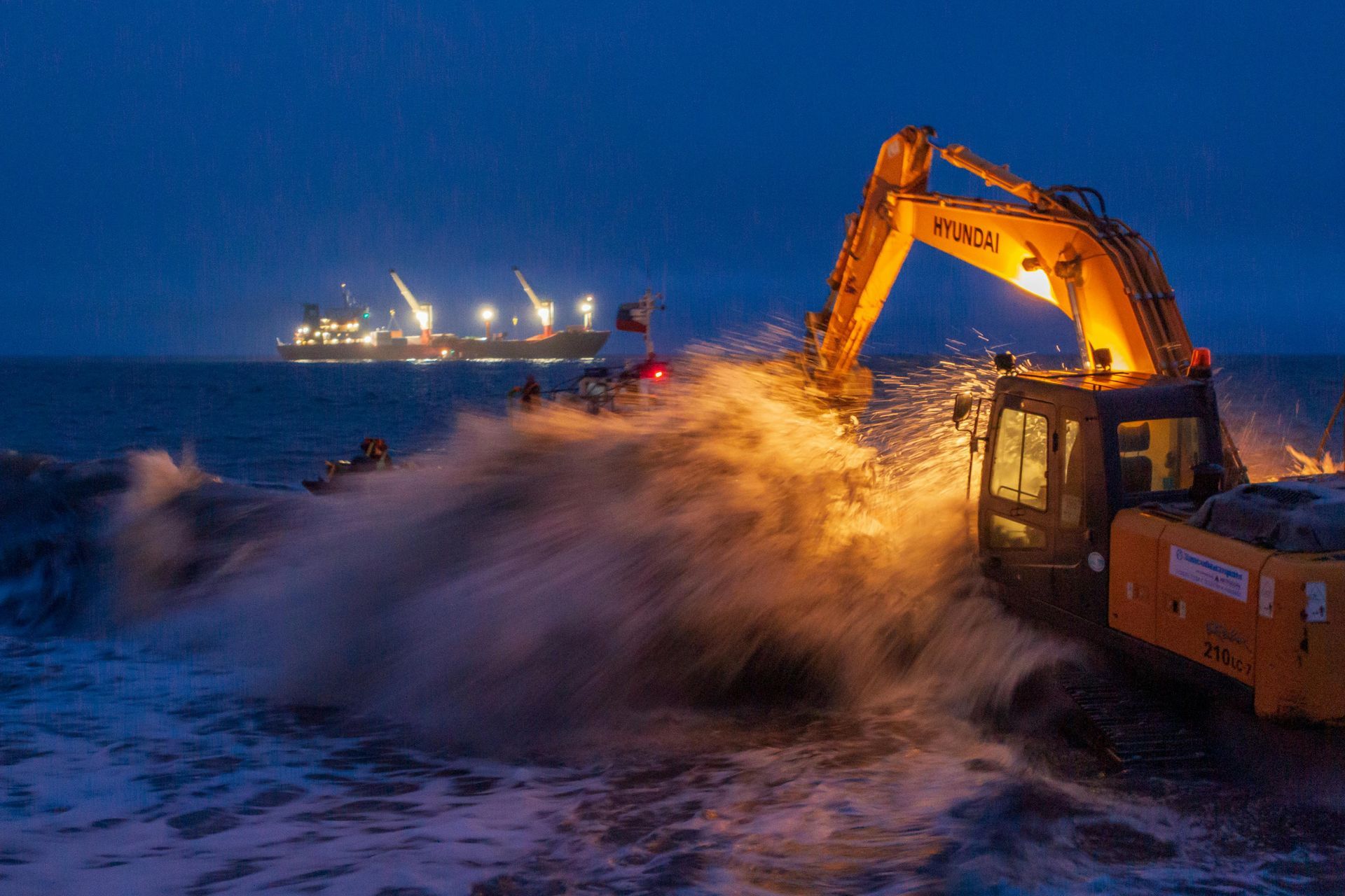 An excavator scoops water in a stormy ocean, illuminated by its lights. A large ship is visible in the background at night.