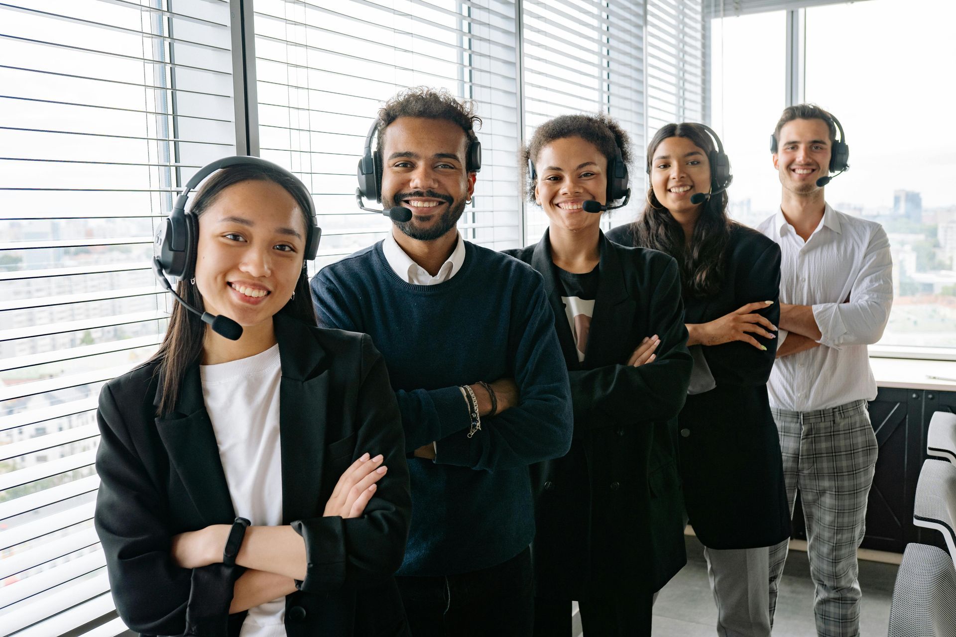 A diverse group of five call center employees with headsets, smiling, arms crossed, standing in front of a window.