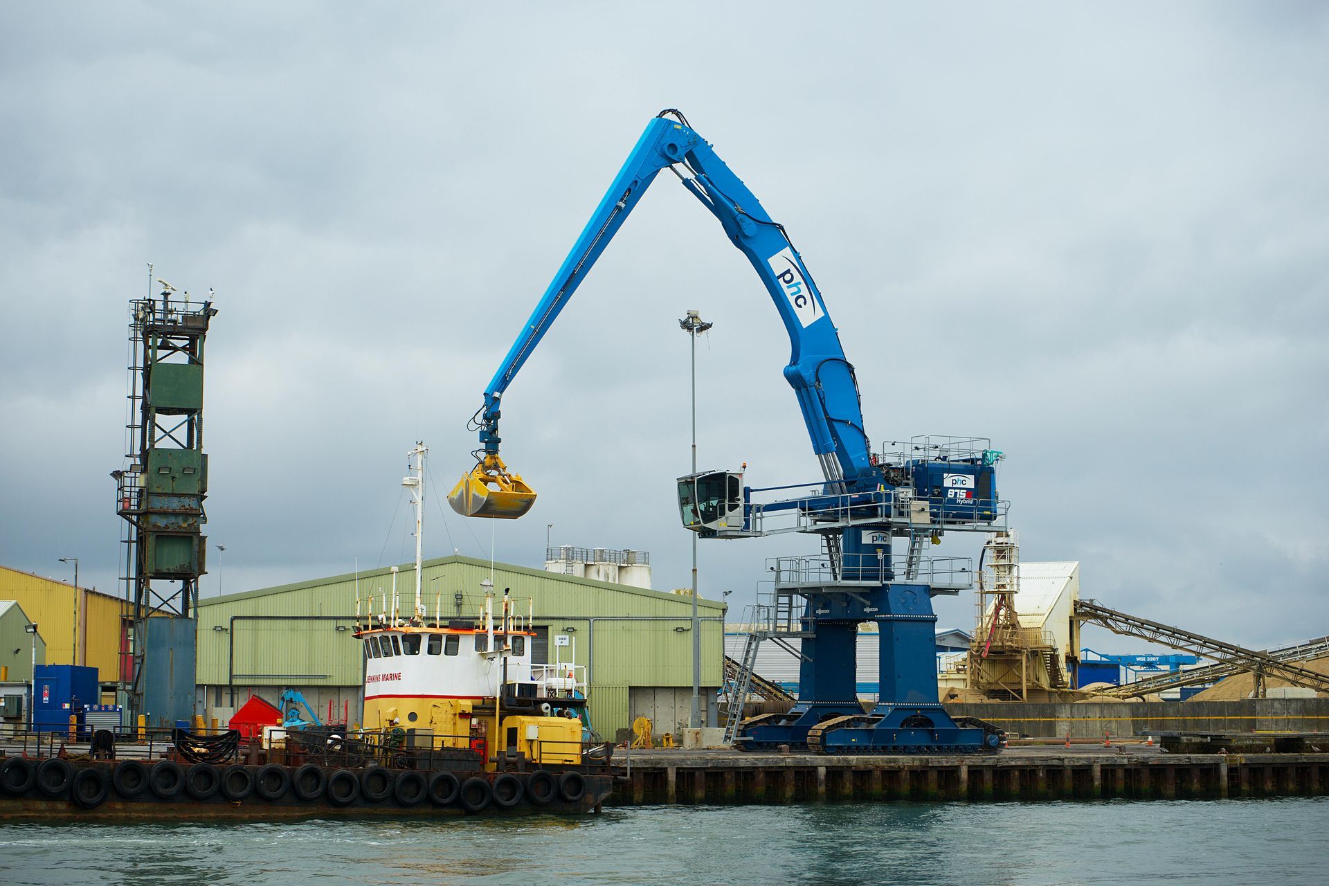 A blue industrial crane with a claw, likely at a port, scoops materials near a tugboat and other structures. Cloudy sky.