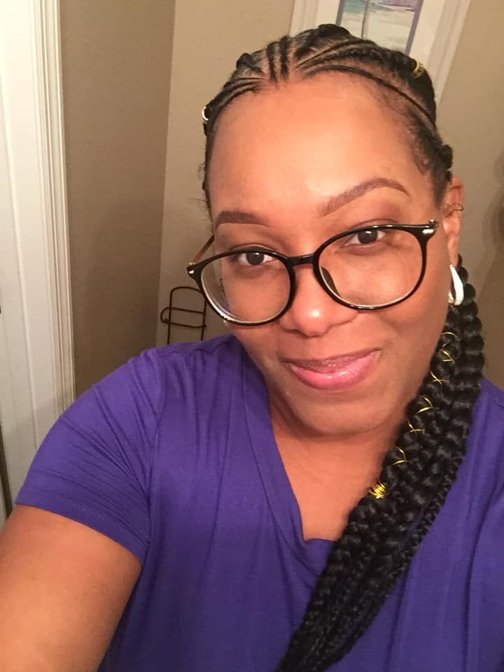 Woman with glasses and braided hair smiles at the camera, wearing a purple shirt, in a bathroom setting.